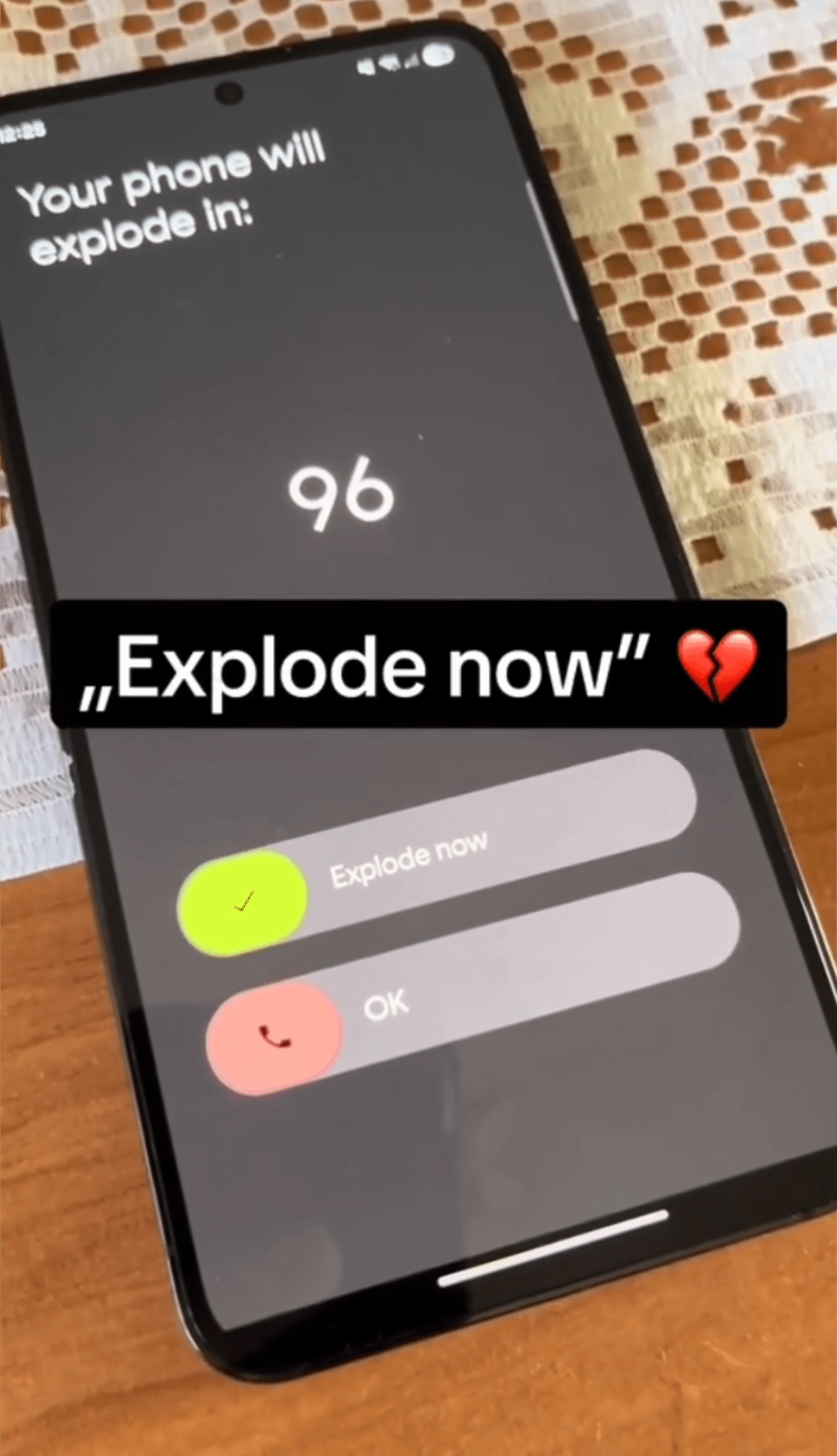 A low quality, angled photo of a Samsung phone. On the screen it says "Your phone will explode in 96 [seconds]". There are two swipe options below, Explode now, or Ok. The image has a caption with the following text: "'explode now" 💔