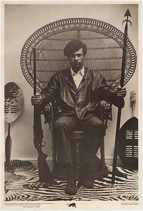 old photography of Huey P. Newton sitting in a chair holding a rifle in one hand and a traditional spear in the other