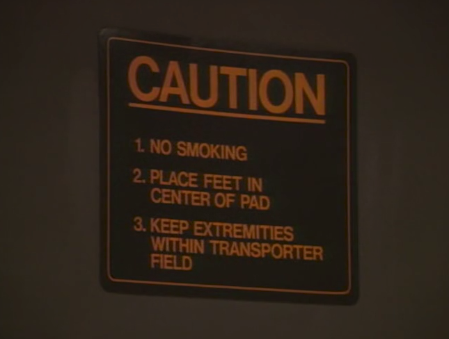 screencap of a sign that says CAUTION above a numbered list: 1. NO SMOKING, 2. PLACE FEED IN CENTER OF PAD, 3. KEEP EXTREMITIES WITHIN TRANSPORTER FIELD