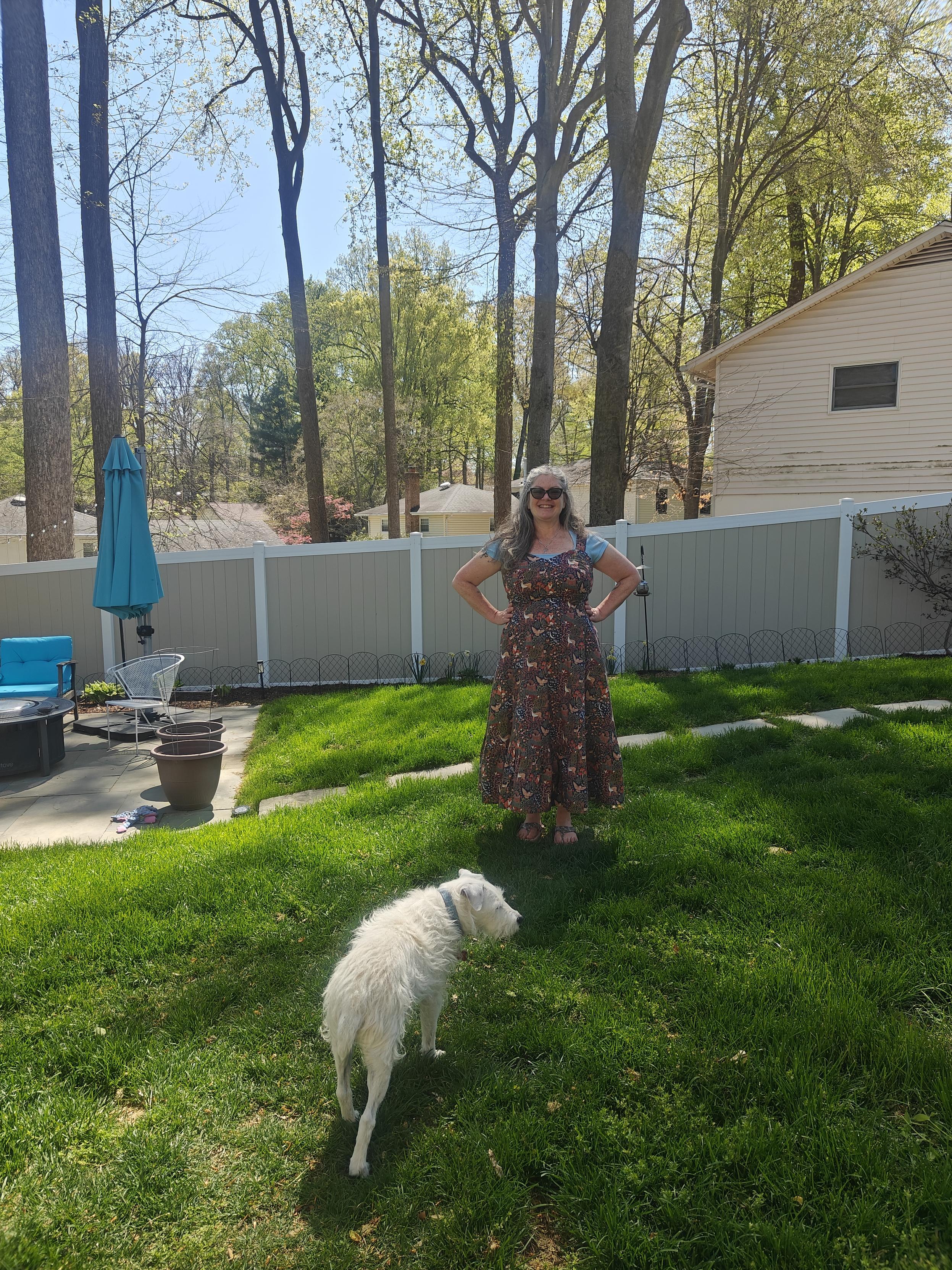 I am standing in my back yard, on the grass. Behind me is a patio with furniture. I am wearing a strappy dress with sweetheart neckline and ruffled hem on the A line skirt, over a pale blue tee. The print on the dress is woodland animals (fox, deer, owl, squirrel, moths) and trees on a navy background 