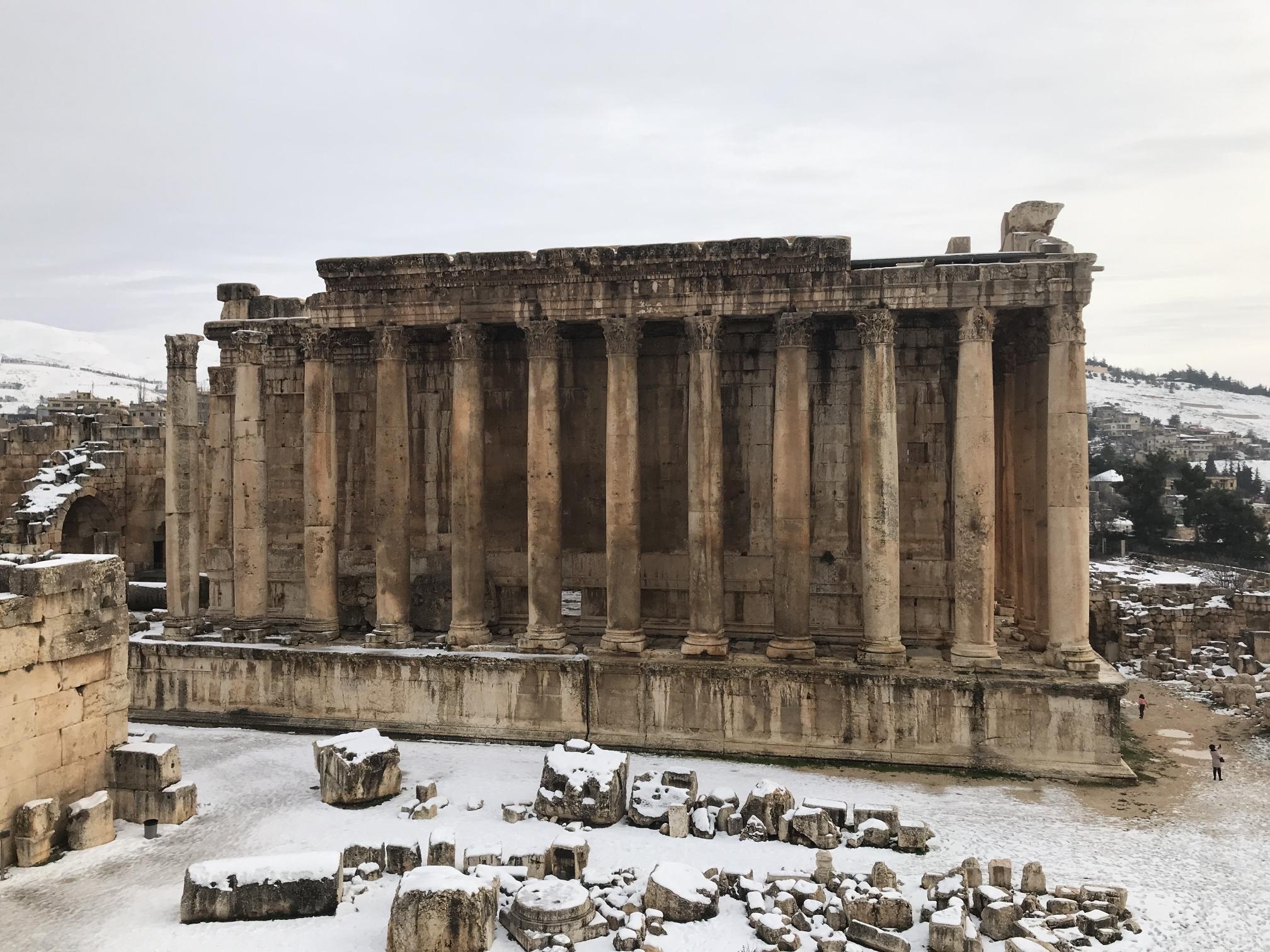 One of the long sides of the temple, roughly 20 meters tall, 60 meters long, and composed of a stone foundation supporting 15 columns, with a solid stone wall behind the columns. The surrounding landscape is littered with ruins and covered with a dusting of snow. Modern buildings are visible in the background.
